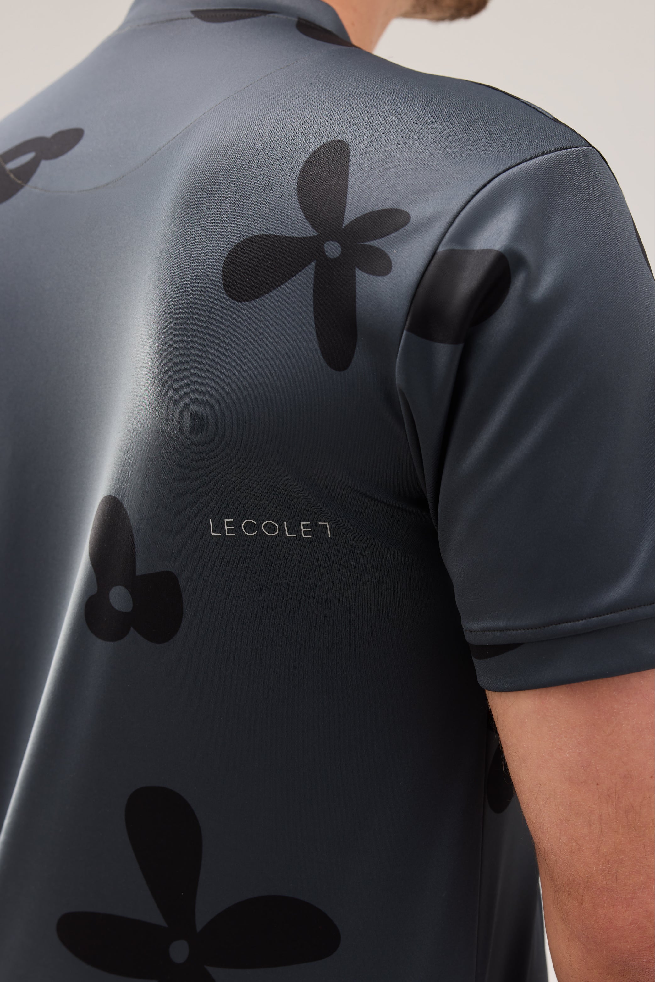 Lecolet SEED golf polo Night Bloom. Dark grey golf polo with darker flower design. Detail white lecolet logo below right scapula.